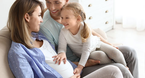 Pregnant woman sat on a sofa with her husband and daughter. Her daughter is touching her tummy
